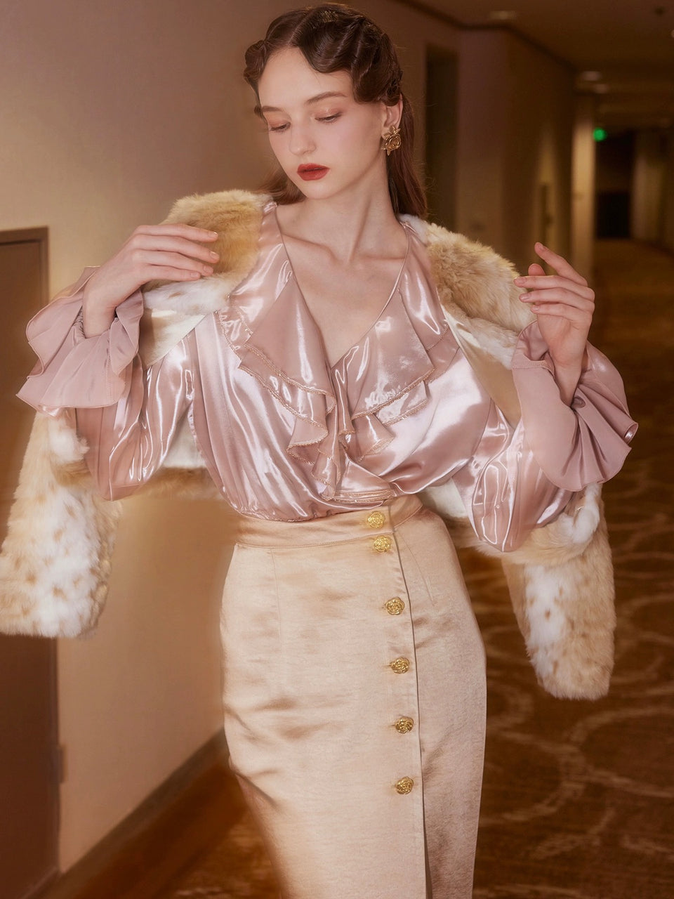 A woman wearing a peach colored, satin two-piece outfit set with a shirt and skirt. The outfit features a V-shaped neckline, sleeveless top, and a skirt with button details.