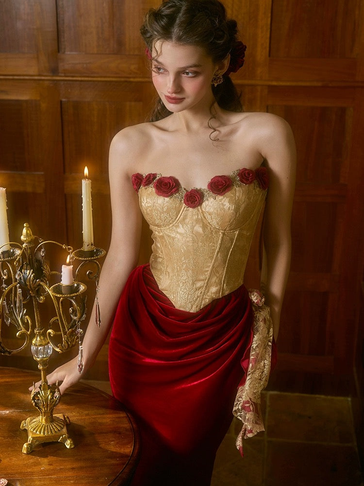 A woman wearing a luxurious golden dress with rose lace details and a red velvet skirt, standing near a candle holder.