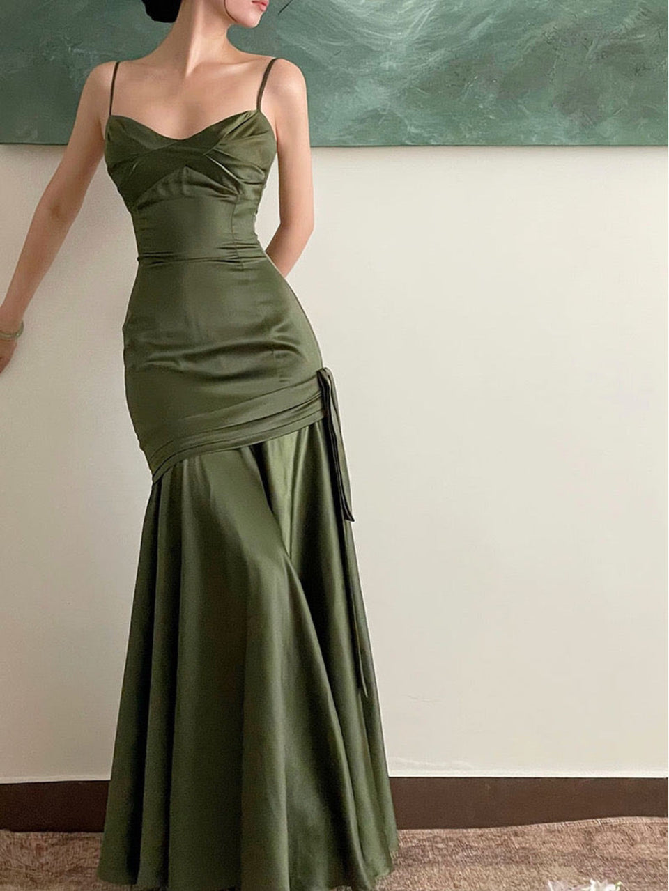 A woman wearing an olive green Arabesque dress with a sweetheart neckline and spaghetti straps, featuring a fitted bodice and a flared skirt with asymmetrical detail.