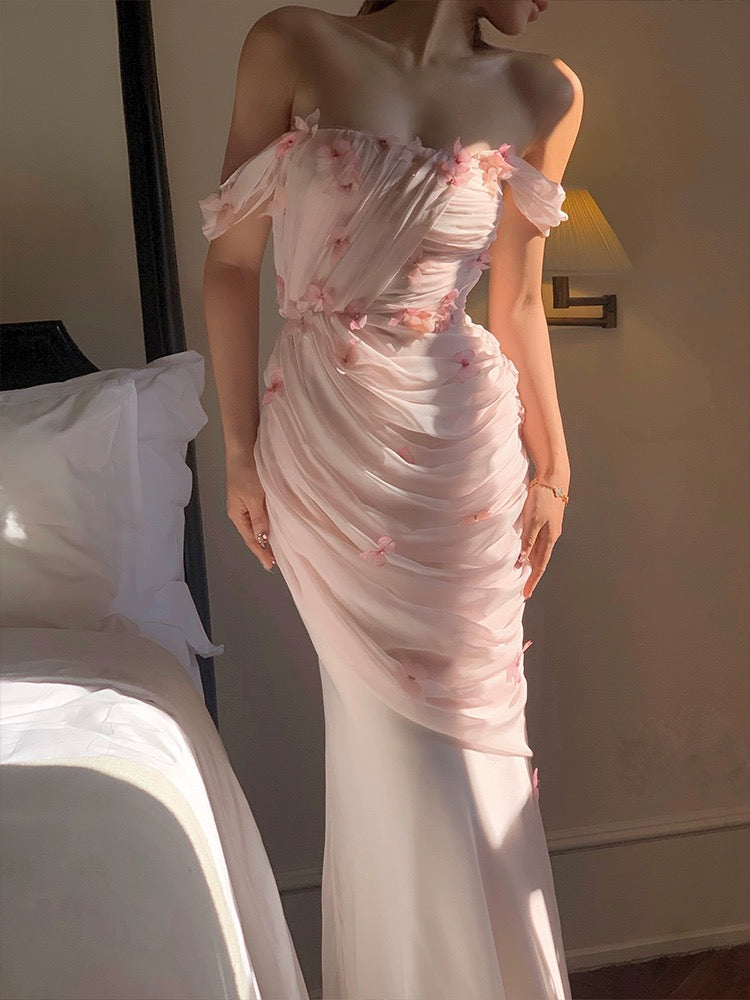 A woman in a formal white dress with pink butterfly embellishments and a flowing silhouette, standing in a room with a bed and a lamp in the background.