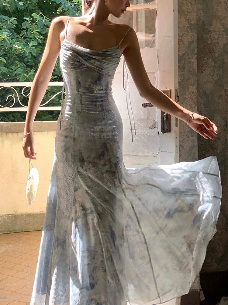 A woman wearing a long, blue, floral dress with a fitted silhouette and sequin detailing, standing in a room with a white door and greenery in the background.