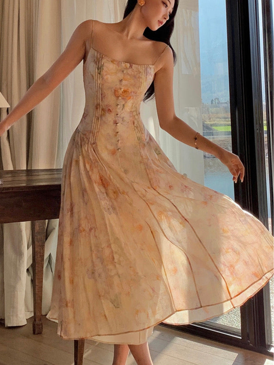 A woman wearing a floral Annesonia dress in a creamy apricot hue with a cinched waist and A-line silhouette, featuring hand-pleated details and a sculpted button at the front.